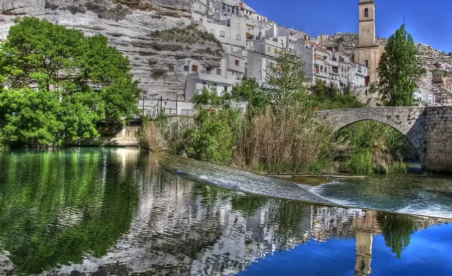 vista panorámica de alcala del júcar con sus casas blancas en la ladera, el puente de piedra y el río júcar reflejando el paisaje